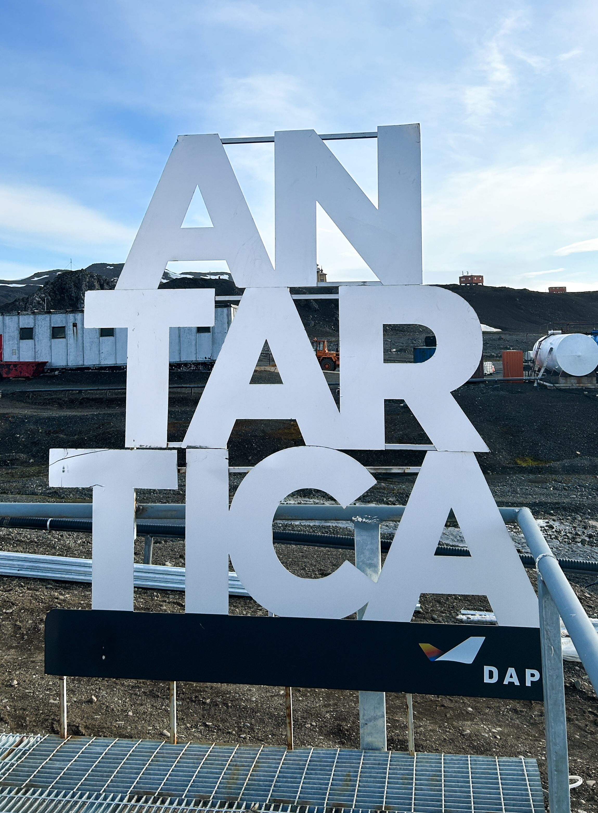 Posted at the DAP terminal on King George Island, large three line sign that spells Antarctica in large white block letters. Behind it brown mountainous landscape.