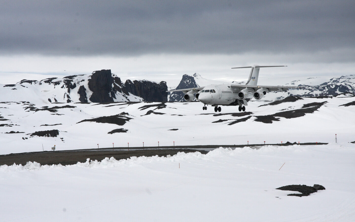 gravel air strip on King George Island, Antarctica. A white airplane is about to touchdown, surrounded by snow-covered mountain range.