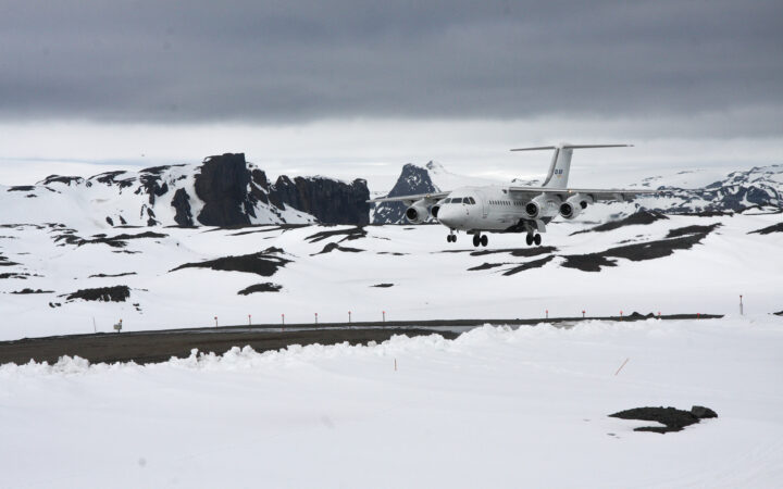 gravel air strip on King George Island, Antarctica. A white airplane is about to touchdown, surrounded by snow-covered mountain range.