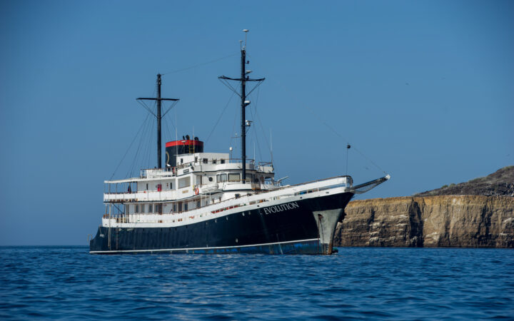 Small ship cruise Evolution anchored off the shore in the Galapagos on sunny, cloud-free day with a rock formation in the distance.