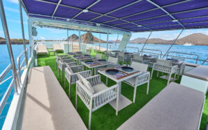 Eight tables set for outdoor dining under a blue awning near sun-soaked chaise loungers aboard Monserrat boat in Galapagos.