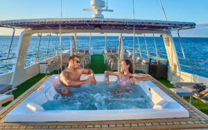 Couple enjoying Jacuzzi on the top deck of small cruise Monserrat on a sunny afternoon. Green turf under outdoor dining area behind them.