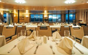 Dining room with white-tablecloth & napkin settings, modern chairs & view windows aboard the Heritage Adventurer ship.