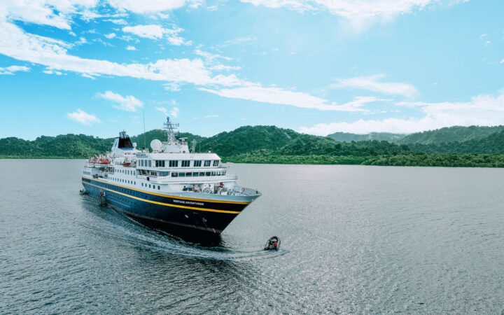 Heritage Adventurer ship with dark blue hull & white upper decks sits in calm sea near green shore as Zodiac boat cruises by bow.