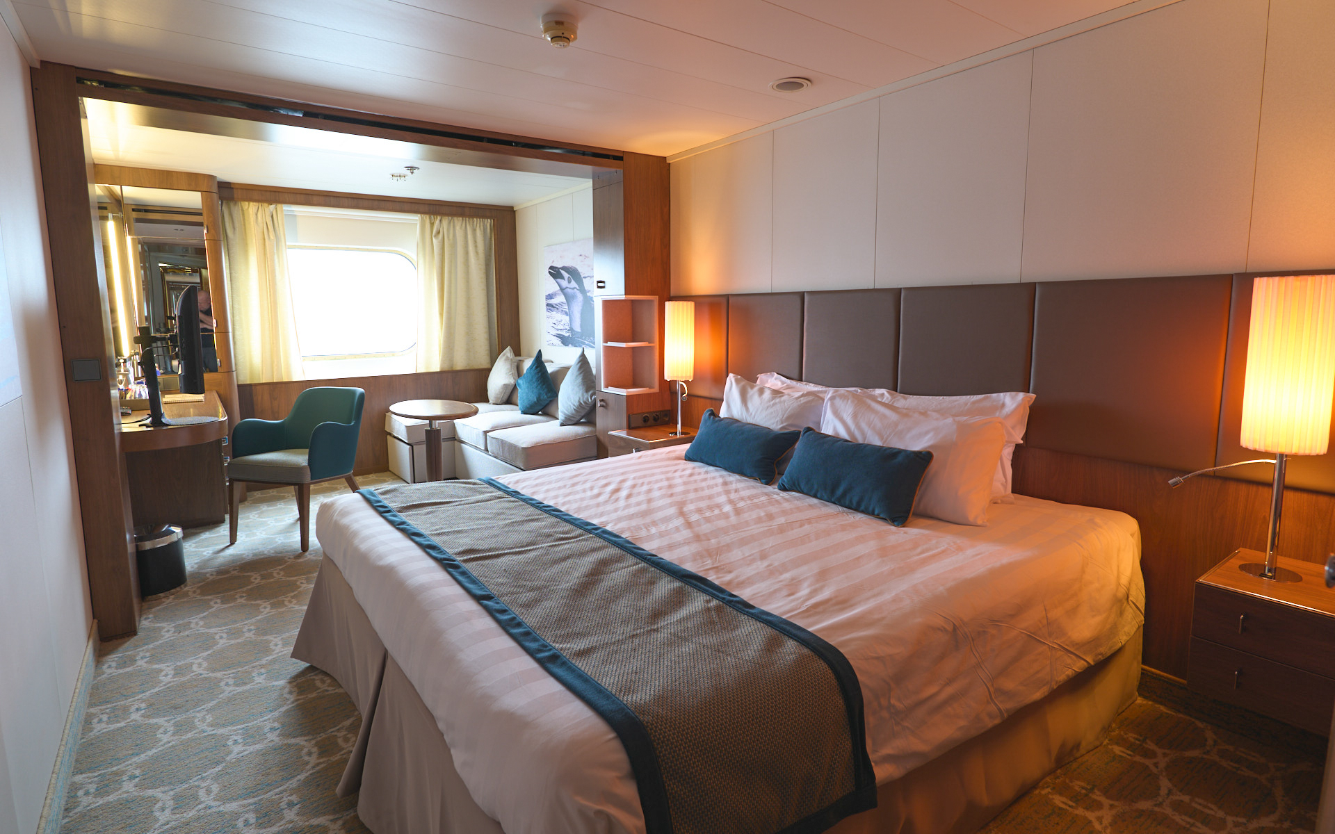 Superior Single on Heritage Adventurer ship with king bed covered in white linens & brown bed runner. Tan couch & window beside it.