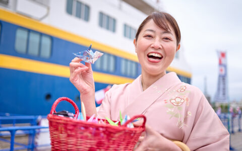 Japanese woman in pink kimono smiles & holds a paper crane from a red basket while standing beside a blue, white & gold ship.