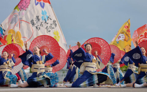 Group of Japanese women in blue kimonos with red parasols perform a dance outside under colorful large flags in the sun.