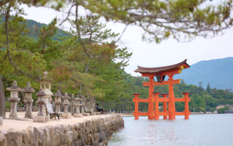 Wooden red torii gate with Japanese symbols sits in water beside stone shoreline backed by green forest in Miyajima, Japan.