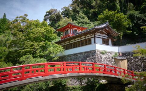 Temple in red, white & brown sits tucked into a hillside by a red pedestrian bridge, seen on a Japan circumnavigation cruise.