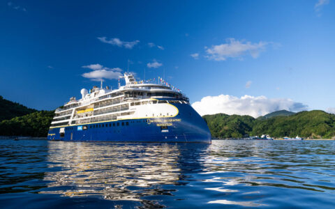 Small expedition ship Nat Geo Resolution with blue, white & gold exterior sits in calm sea offshore of green islands in Japan.