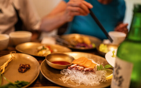 Golden plates & bowls with red sauce, rice noodles & raw seafood sit beside a green beer bottle at a meal in South Korea.