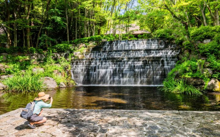 Woman in shorts & sun hat kneels atop stone pavilion to photograph a cascading waterfall among bright green trees in Japan.