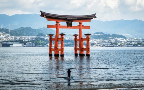 Man walks into shallow water towards towering red wooden torii gate sitting in calm sea backed by a modern city in Japan.