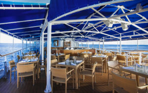 Al fresco dining area on La Pinta small ship with rattan furniture, glass-top tables, blue awning overhead & white structural poles.