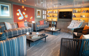 Spacious lounge aboard the La Pinta small ship. 3 striped couches and 3 accent chairs surround 2 coffee tables. Bookshelf in background.