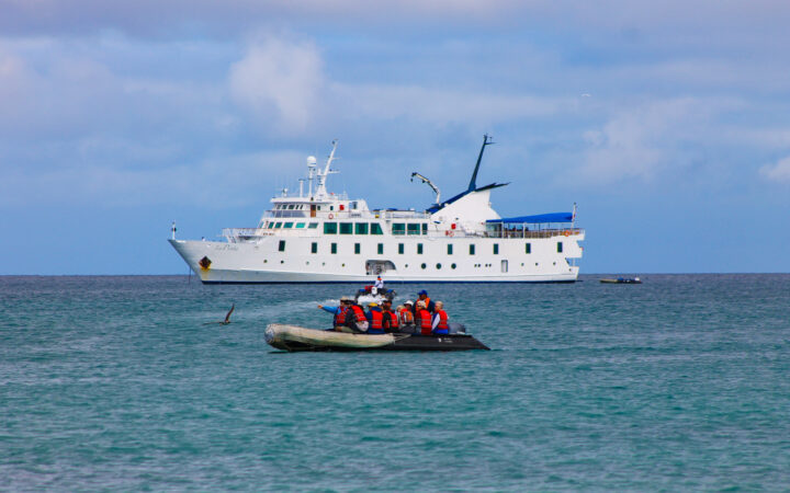 Exterior of the La Pinta small cruise seen on its port side. Zodiac with travelers in orange life vests on water in foreground.