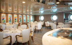Dining area showing 6 tables set for a meal next to large port holes aboard La Pinta small cruise. Reflective ceiling and warm lighting.