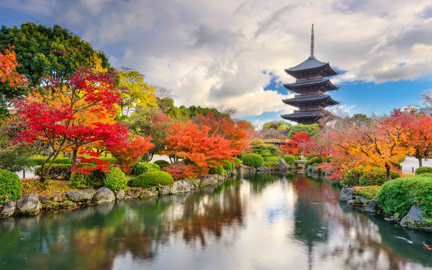 4-level brown pagoda sits by glassy pond, bright maple trees & green shrubs, seen on luxury small ship cruises in Japan.