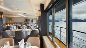 Restaurant on Magellan Explorer ship with grey & tan leather chairs around white table clothed tables & view windows.