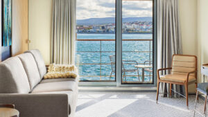 Shared area with grey sofa across from desk, stool & chair in corner overlooking balcony from Grande Suite on Magellan Explorer