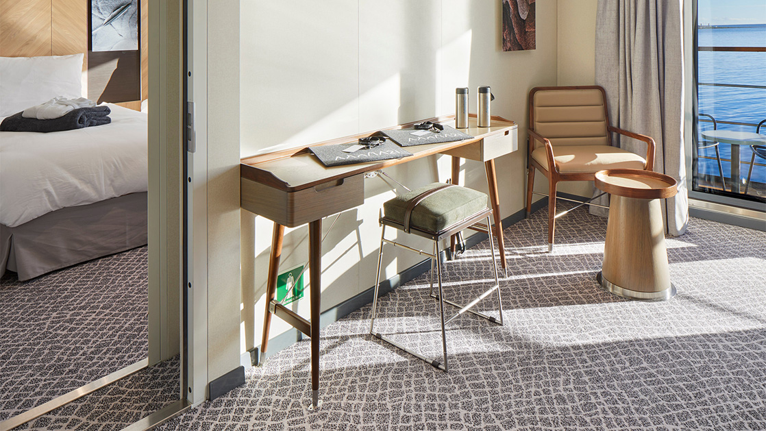 Corner of shared area with desk, cushioned stool & chair with side table overlooking balcony from Grande Suite on Magellan Explorer