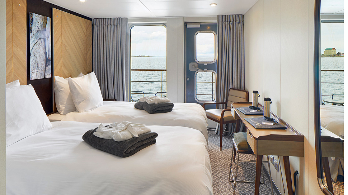 Veranda Cabin on Magellan Explorer with 2 twin beds across from desk with cushioned stool & chair in corner overlooking balcony