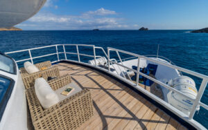 Forward-facing small teak deck with 2 padded chairs facing blue sea & small volcanic islands on Monserrat boat in Galapagos.