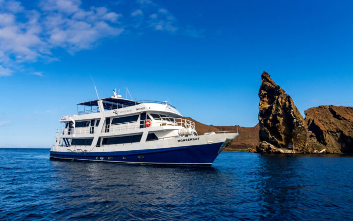 Monserrat boat in Galapagos with dark blue hull, white upper decks & 4 guest decks sitting beside Kicker Rock on a sunny day