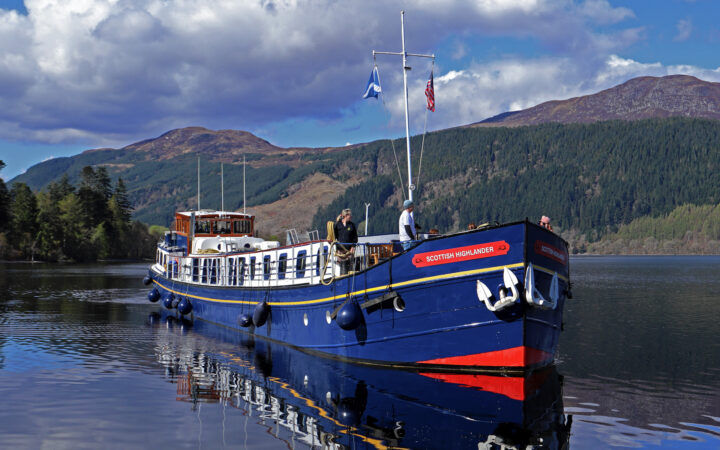 Scottish Highlander hotel barge sitting still in calm water with red-&-blue exterior & on a sunny day in Scotland.