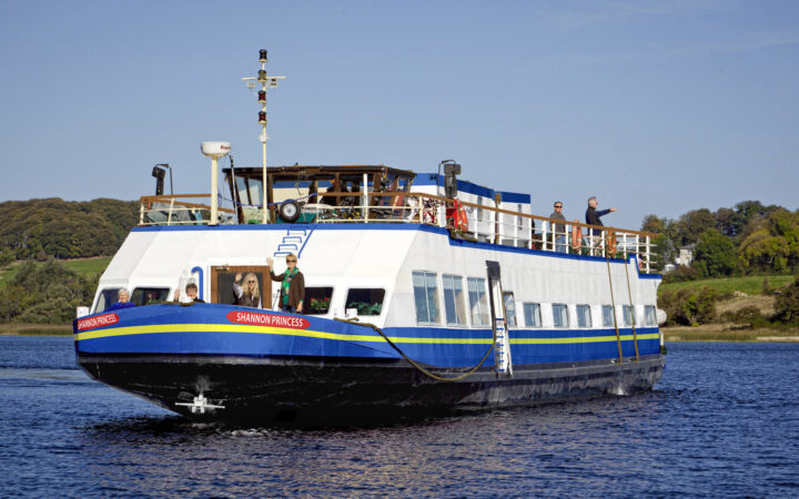 Bow view of Shannon Princess barge with blue hull & 2 white upper decks as she cruises through calm sea in the sun.