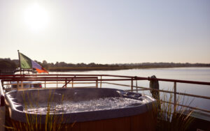 View of hot tub on upper deck aboard Shannon Princess barge cruising Ireland's River Shannon. Ocean view, flag of Ireland, plants in foreground.