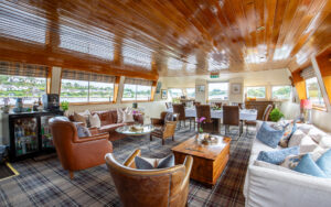 Common area with soft white & tan leather couches, wooden coffee tables, carpets, wooden ceiling, large view windows & 10-guest wooden dining table aboard Shannon Princess barge.