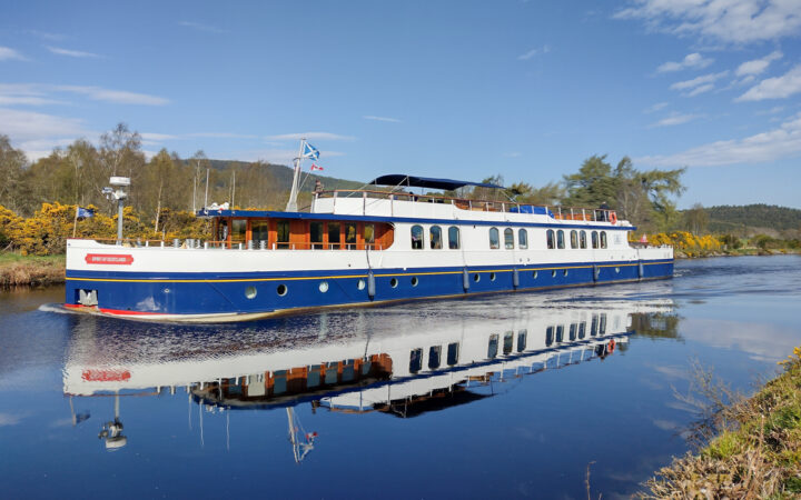 Spirit of Scotland hotel barge going through a Sottish loch on a sunny day with a reflection in the water below.