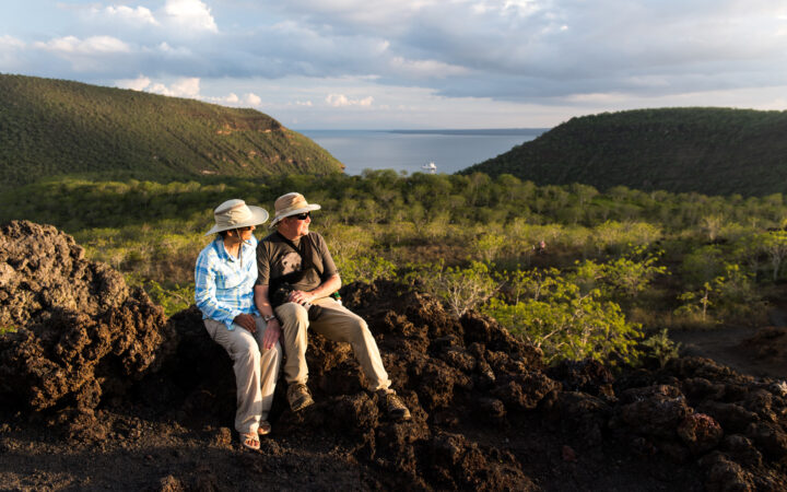 A male and female traveler sit atop lava rock at Tagus Cove in the Galapagos Islands with green trees and the ocean in the distance below them.