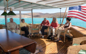 Travelers relaxing on the back deck of small ship Westward. Ocean view behind passengers with an American flag on a sunny day.