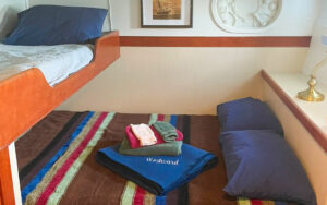 Cabin 1 aboard small ship Westward. Bunk beds with brown, blue, red and green striped comforter. Dark blue pillows and towels displayed on bed.