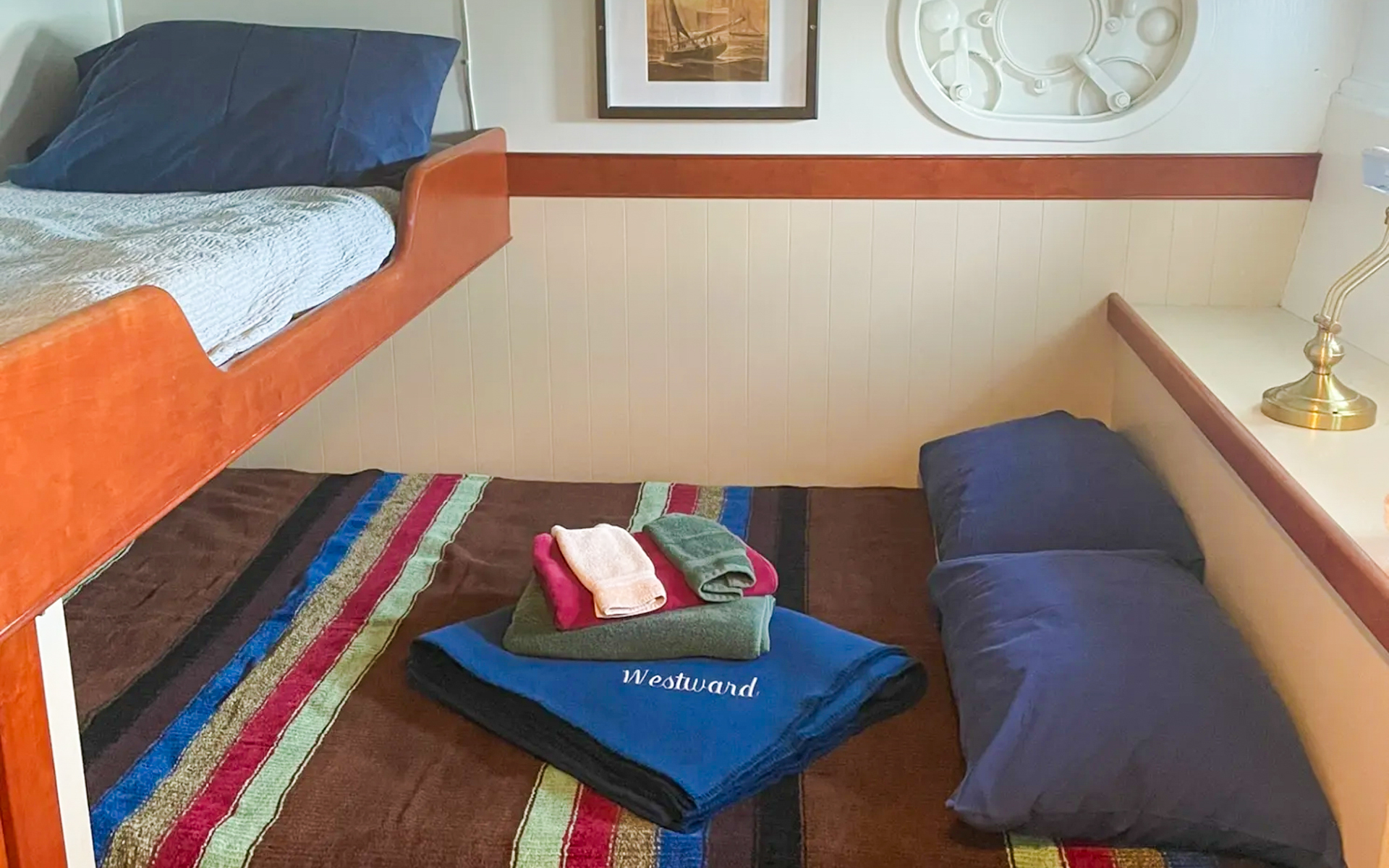 Cabin 1 aboard small ship Westward. Bunk beds with brown, blue, red and green striped comforter. Dark blue pillows and towels displayed on bed.