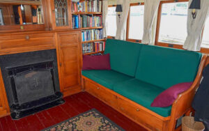 Interior living room area aboard historic yacht Westward. Green cushions on wooden couch next to black iron fireplace and book shelf.