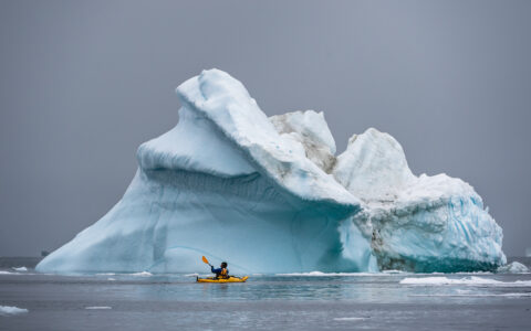 Kayaker passes by large iceberg floating in the middle of the water in a yellow kayak with winter gear during Arctic Wildlife Sarfari