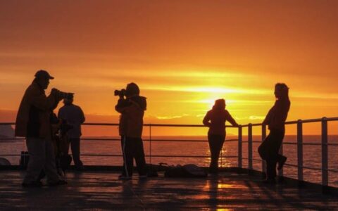 Passengers from the expedition cruise classic antarctica look over the railing of the ship while the sun sets in the background