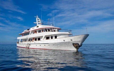 A luxury yacht, the Passion motors over calm, blue waters with the sky and clouds in the background at the Galapagos islands