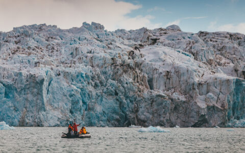 Group of travelers in orange jackets take skiff ride in front of massive arctic glacier during Spitsbergen Highlights small ship cruise