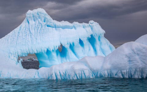 Large blue & white iceberg rises out of calm sea under a dark sky in Antarctica, with a hole showing a small ship nearby.