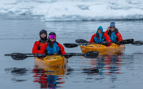 Bundled up tandem kayakers in yellow boats pause for a photo while paddling glassy waters among icebergs in Antarctica.