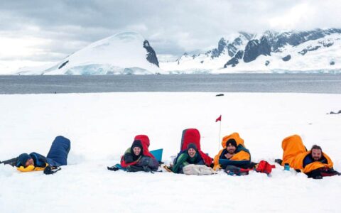 travelers lay on the ground in sleeping bags in antarctica with the ocean behind them and mountains in the distance