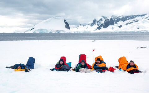 travelers lay on the ground in sleeping bags in antarctica with the ocean behind them and mountains in the distance