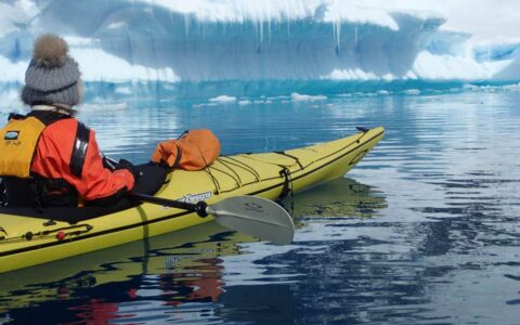 adventure traveler in a yellow kayak sits in calm water looking at an iceberg on the antarctic explorer small ship cruise trip