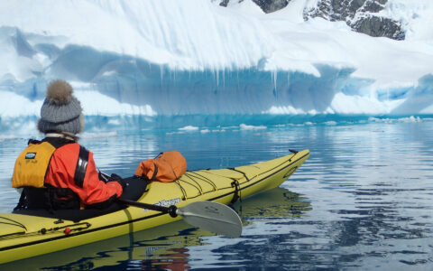adventure traveler in a yellow kayak sits in calm water looking at an iceberg on the antarctic explorer small ship cruise trip