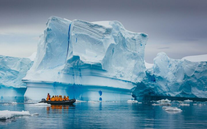 adventure travelers wearing yellow jackets in a zodiac skiff cruise in front of a large iceberg on an overcast day in antarctica