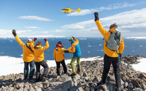 Group of guests in yellow coats stand atop rocky summit & wave to yellow helicopter on a sunny day in Antarctica.